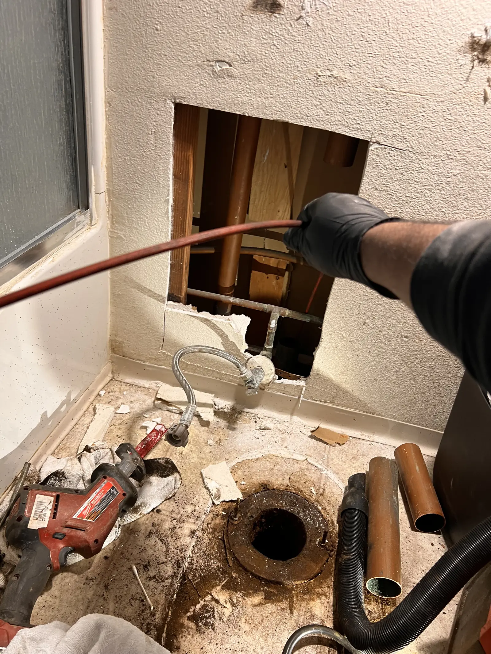A gloved hand feeding a drain cable into an opened floor drain beside a wall cut-out