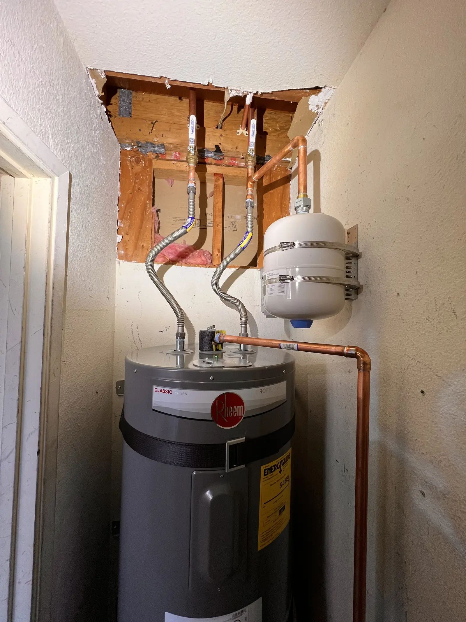 An electric water heater in a garage with an expansion tank and copper lines above it