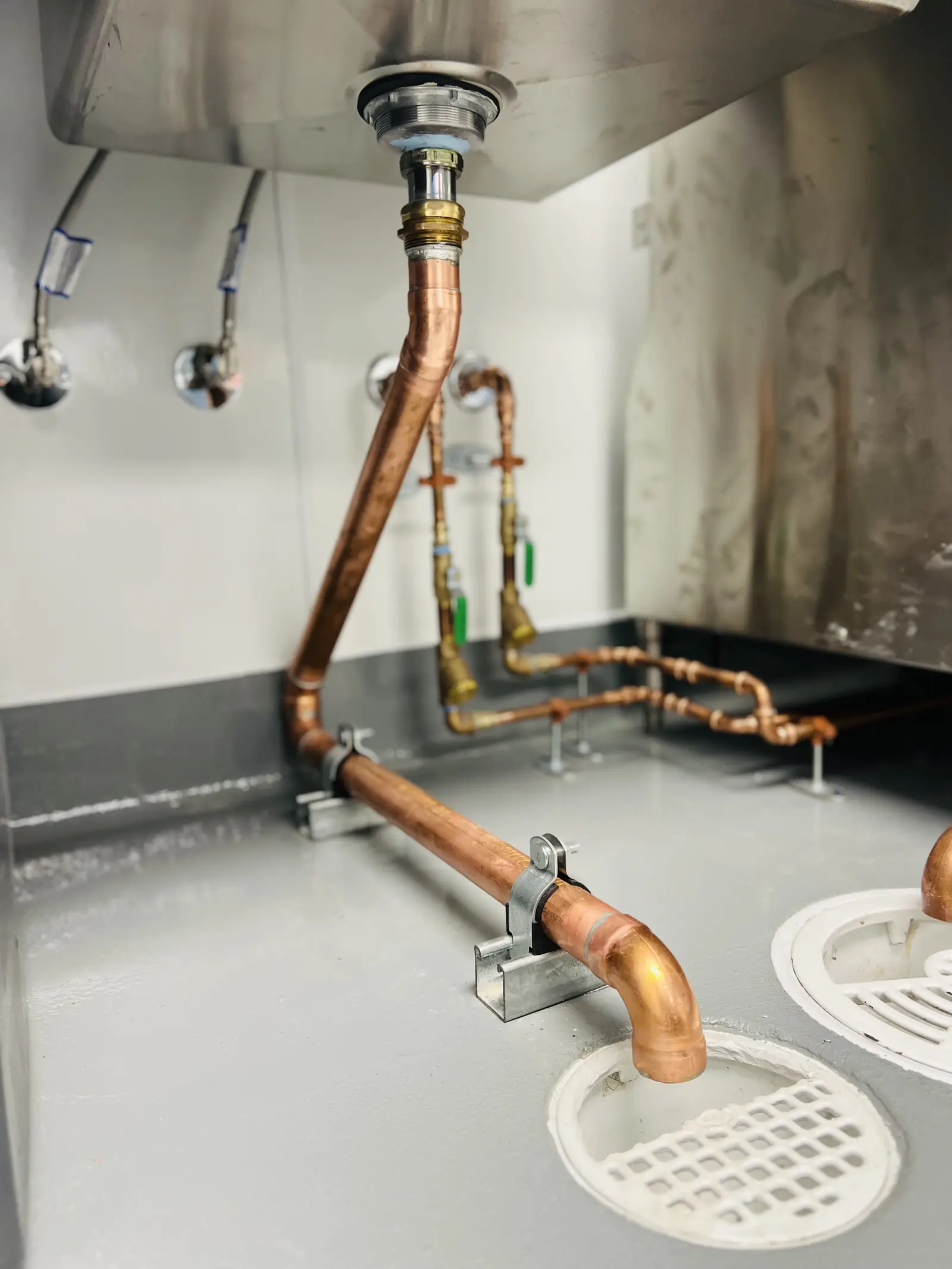 Copper drain lines running from a commercial sink to two white floor drains
