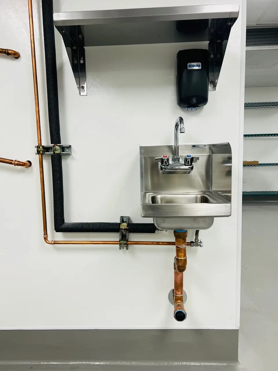 A stainless hand sink on a white wall with copper supply lines and a soap dispenser
