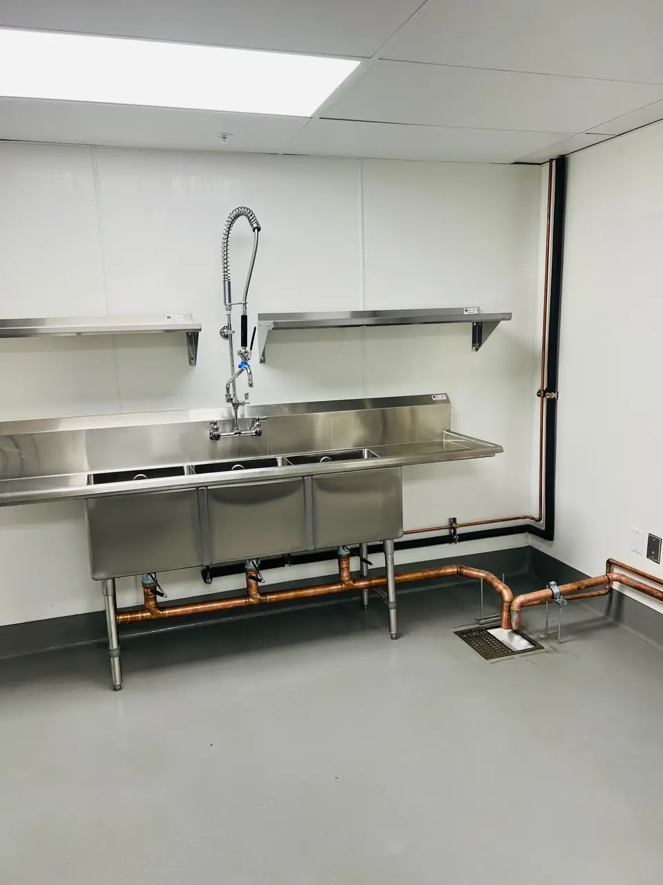 A three-compartment stainless sink with a pre-rinse sprayer and copper lines under it