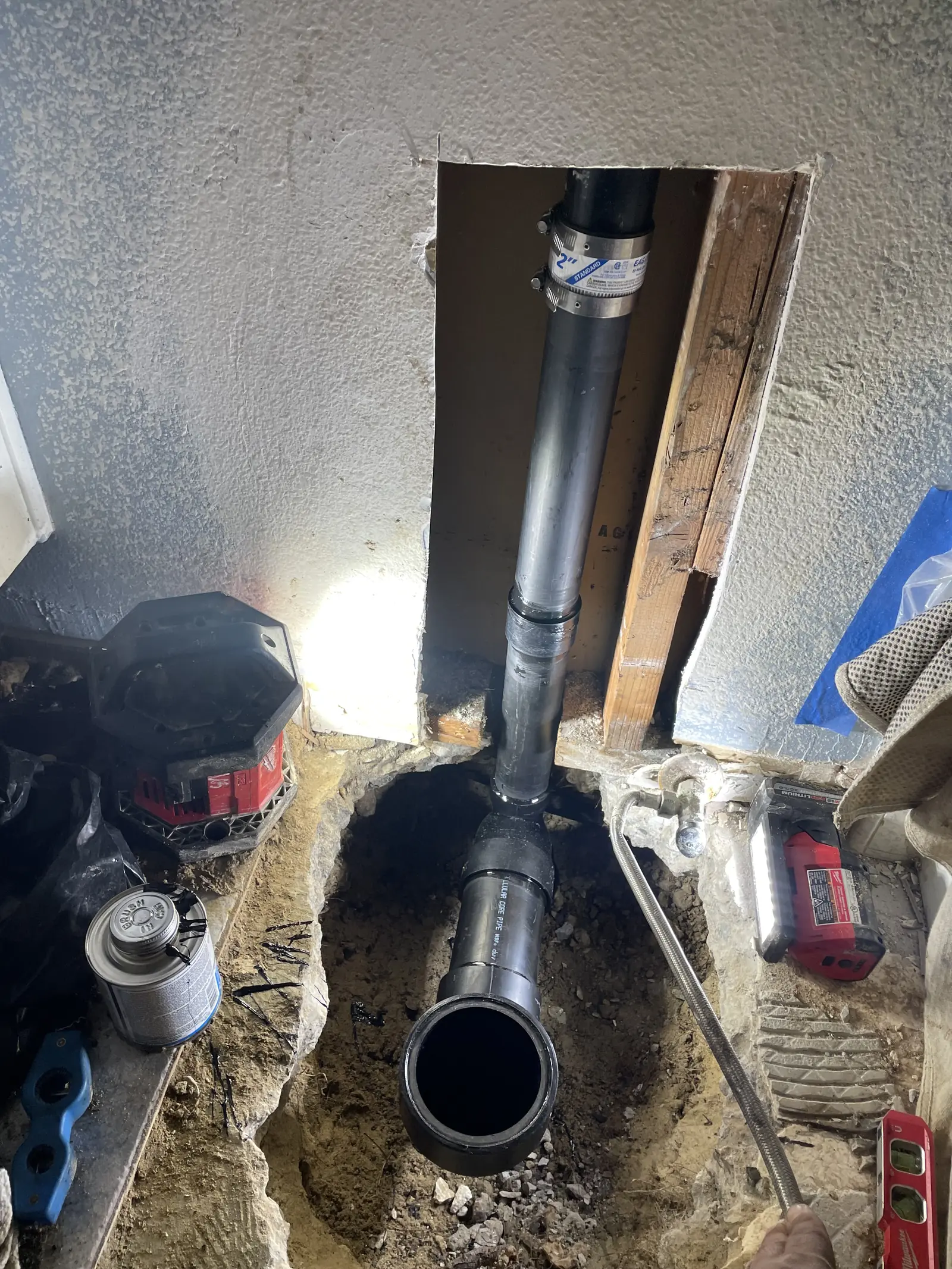 A trench under a house with a black drain pipe rising out of it, tools and a work light beside it