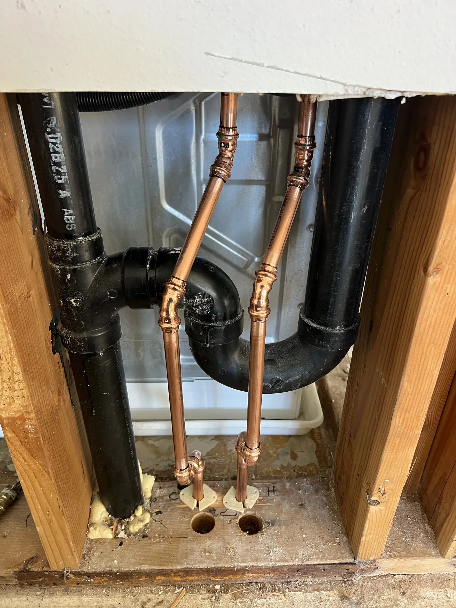 An opened wall showing black drain pipe and two new copper stubs through the floor