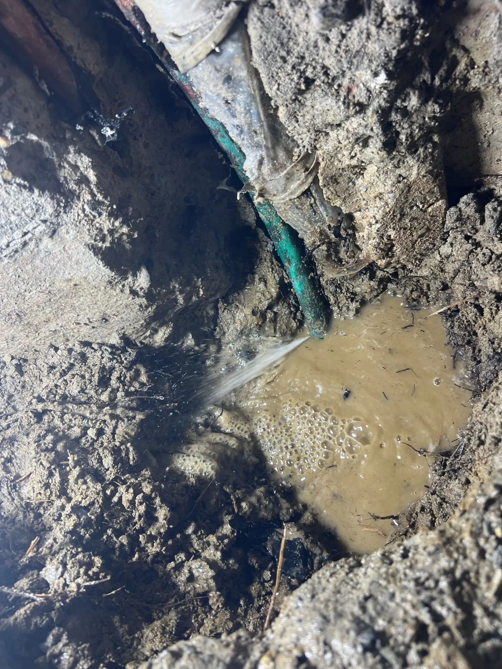 A broken water line in a muddy trench with water spraying from the pipe